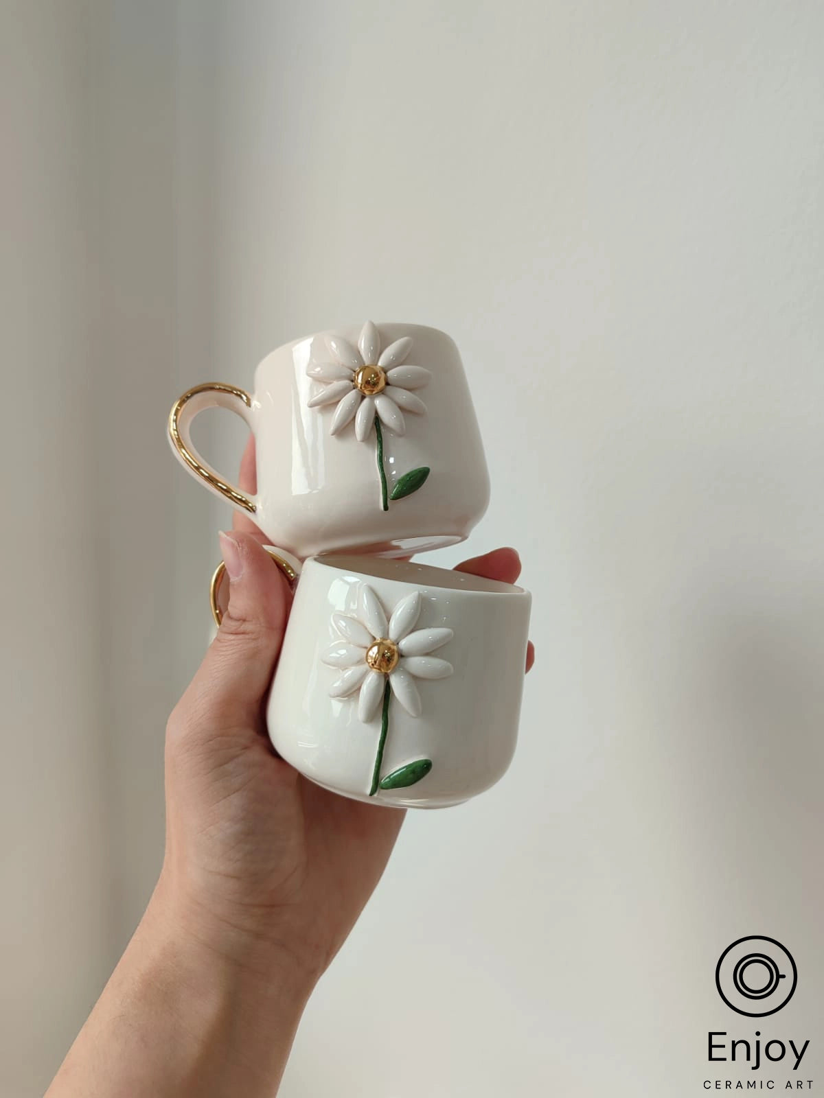 A hand holds two stackable white mugs with gold handles and 3D daisy designs, captured in soft light against a neutral background.