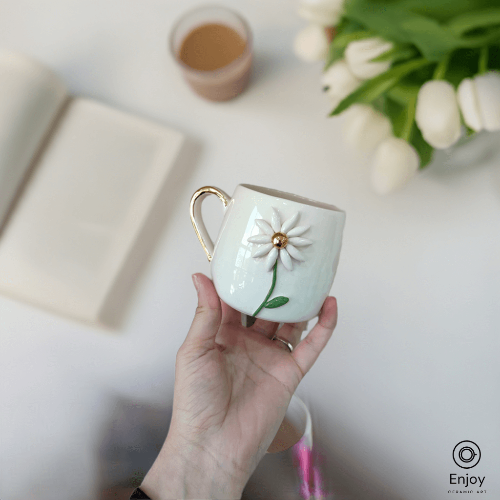 A handmade white ceramic mug with a daisy flower and gold handle is ...