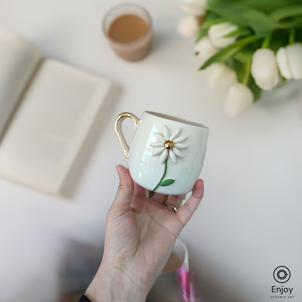 A handmade white ceramic mug with a daisy flower and gold handle is held against a backdrop featuring a cup of tea, an open book, and white tulips.