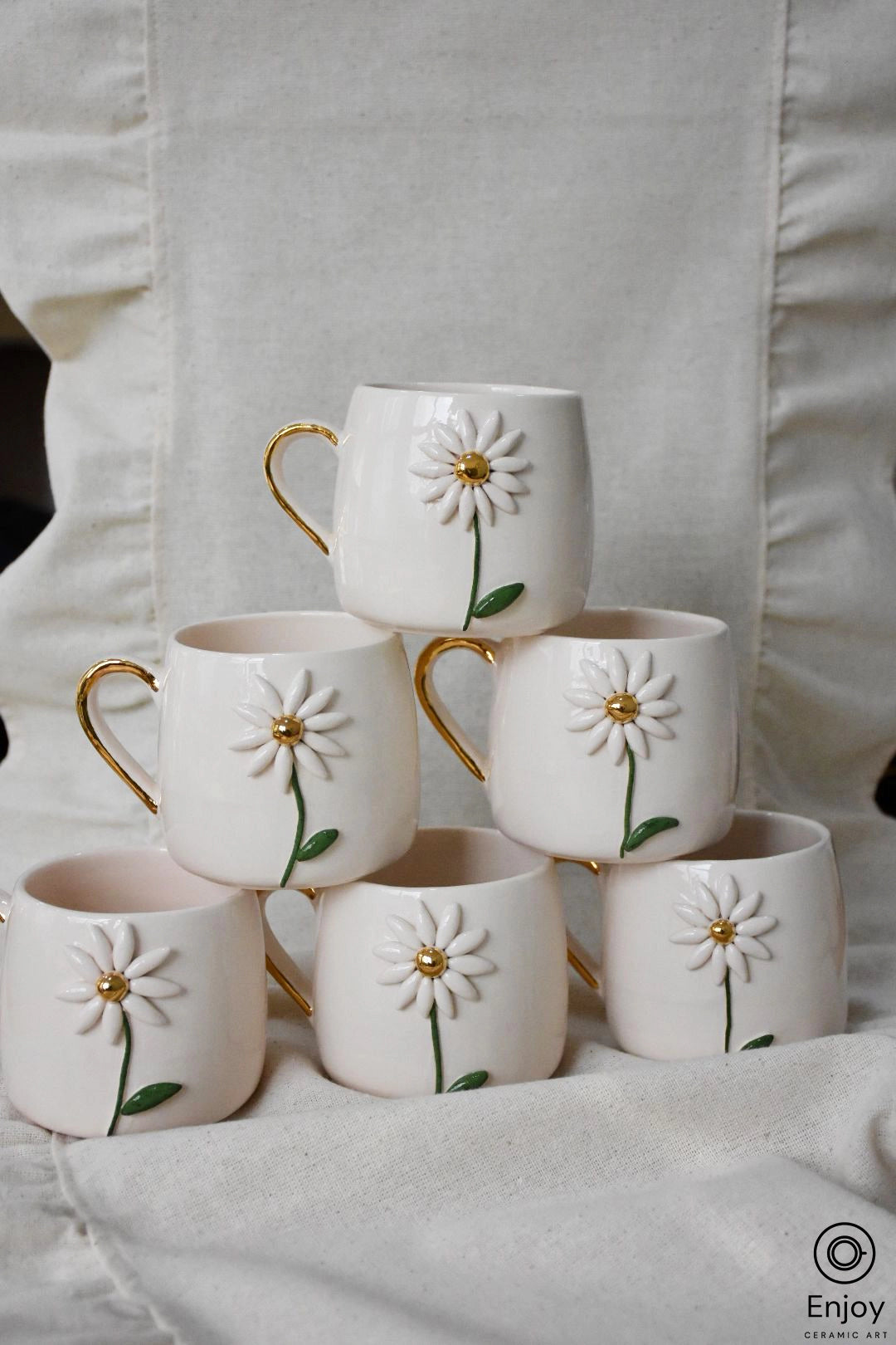 A pyramid of elegant white mugs with relief daisy designs and golden handles, artistically arranged against a linen backdrop.