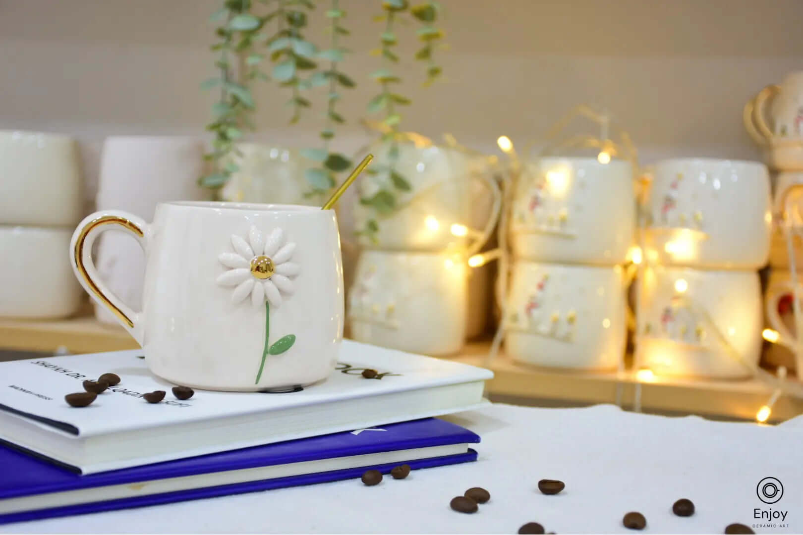 White ceramic mug adorned with a daisy design and gold accents, set on books with coffee beans scattered around, warm lights in the background.