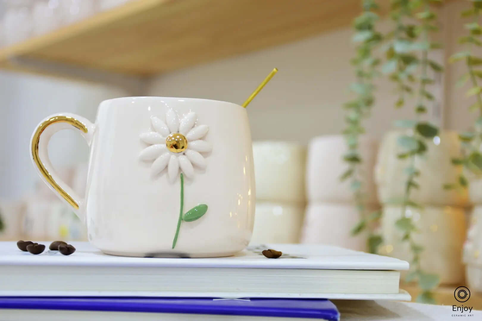 White ceramic mug with a daisy flower design and gold accents, placed on books with coffee beans nearby, green foliage in the background.