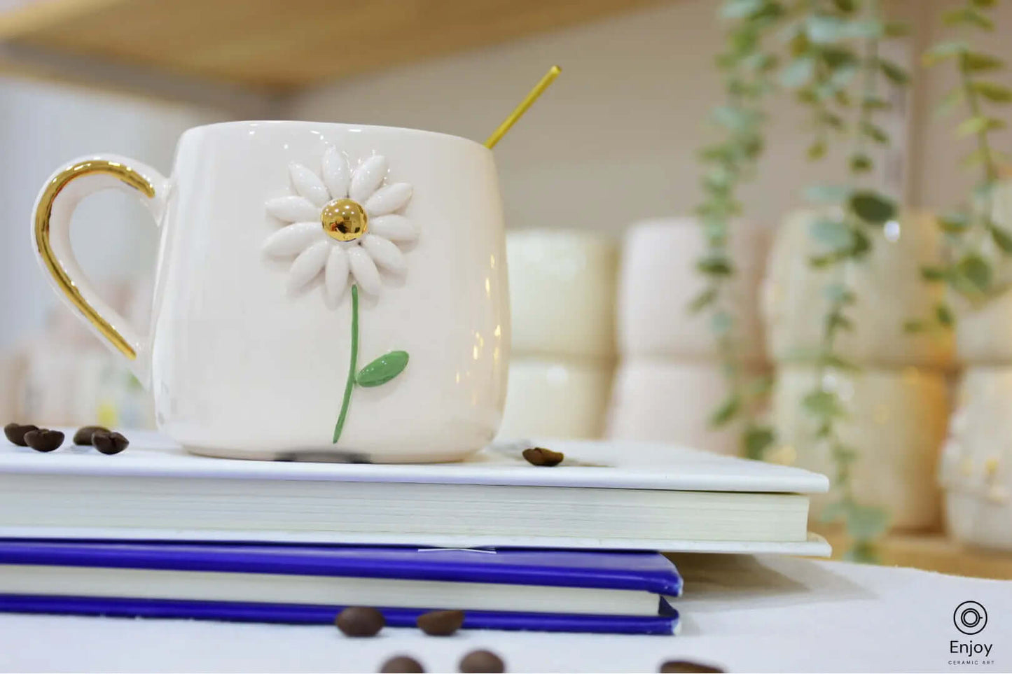 White ceramic mug with a daisy design and gold handle, placed on books with coffee beans scattered, surrounded by greenery in the background.