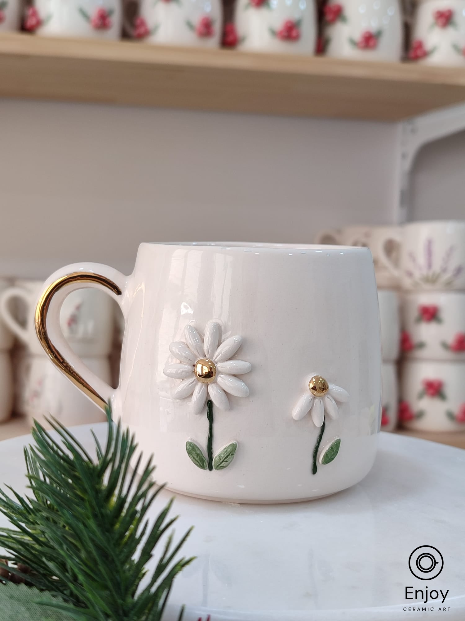 Handmade ceramic mug featuring two delicate daisies with golden centers, accented by a gold handle, displayed on a marble stand with festive greenery.