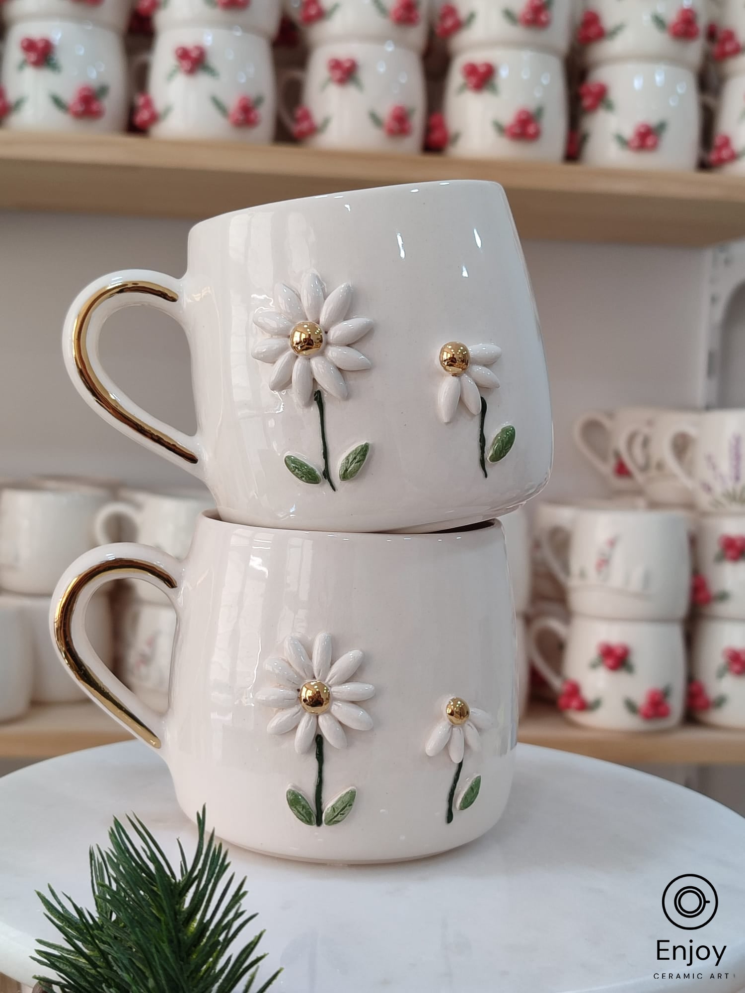 Handmade ceramic mugs featuring two daisies with gold centers and a golden handle, stacked on a marble stand with festive greenery.