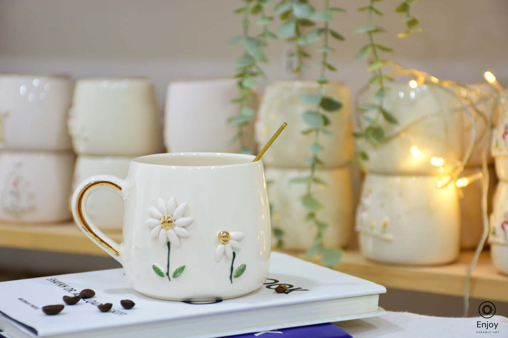 White ceramic mug with two daisies and gold accents, set on stacked books with scattered coffee beans, surrounded by greenery and fairy lights.