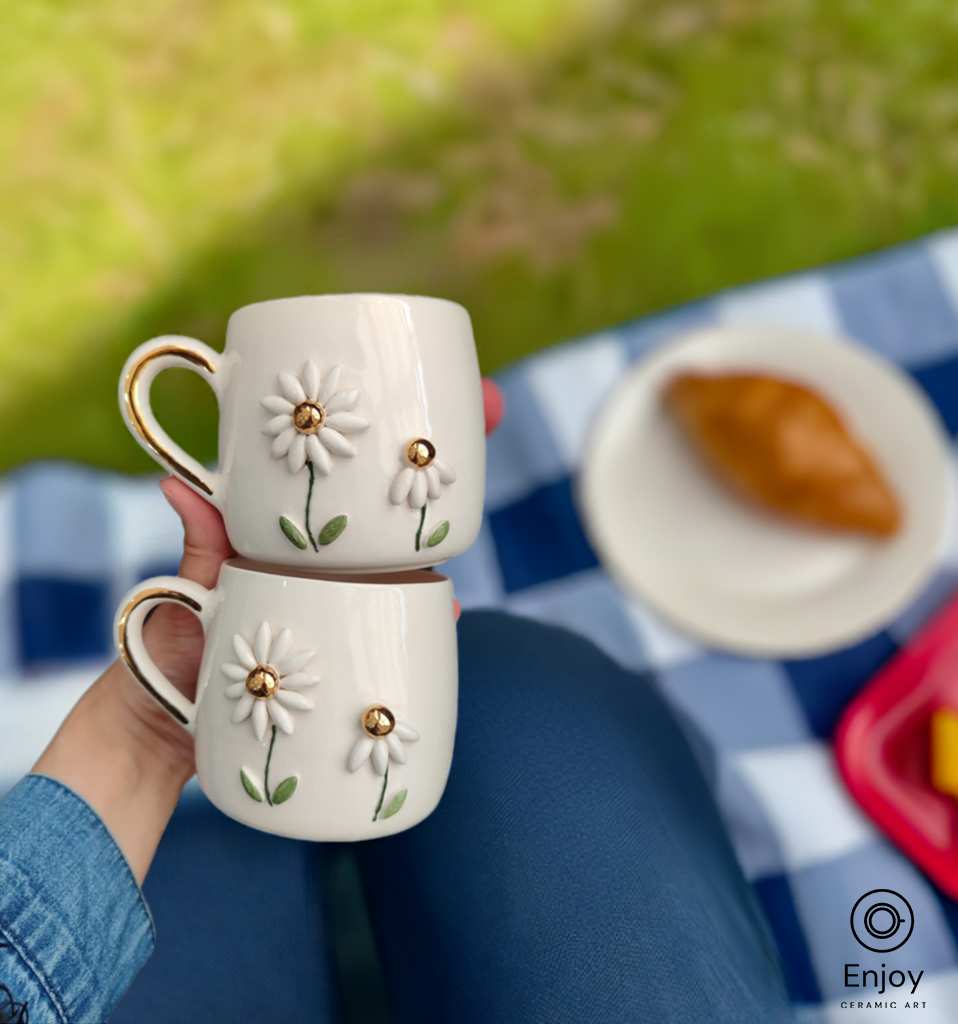 Two handmade daisy mugs with gold accents, stacked in hand, perfect for a picnic backdrop.