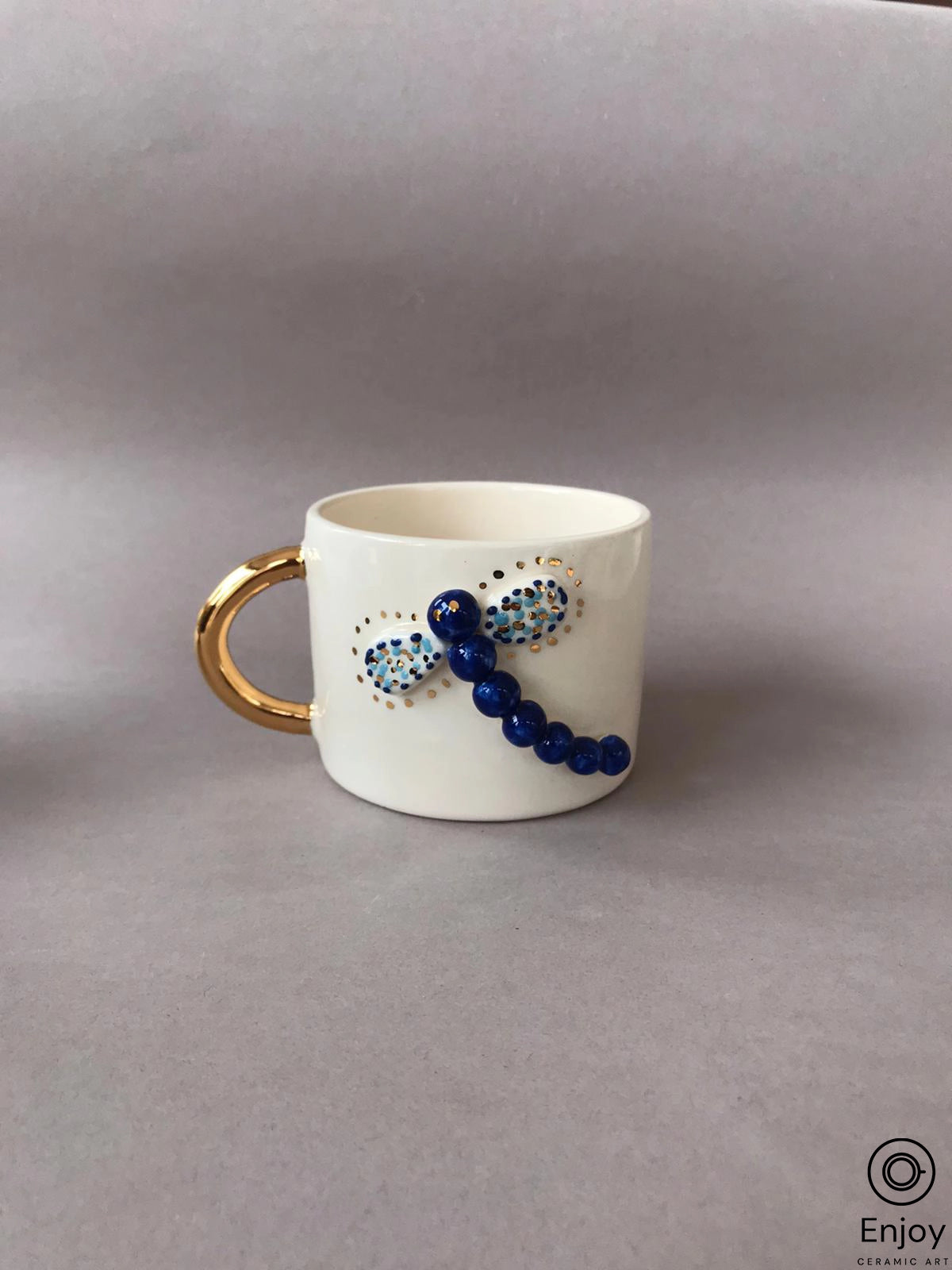 Handcrafted 'Blue Dragonfly Handmade Ceramic Coffee Mug' with a gold handle, displayed on a lavender background for a simple and elegant presentation.