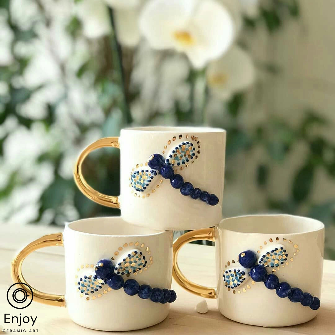 "Three 'Blue Dragonfly Handmade Ceramic Coffee Mugs' with gold handles, elegantly displayed against a backdrop of white orchids. The mugs feature a blue beaded dragonfly design, adding a touch of artisanal charm."