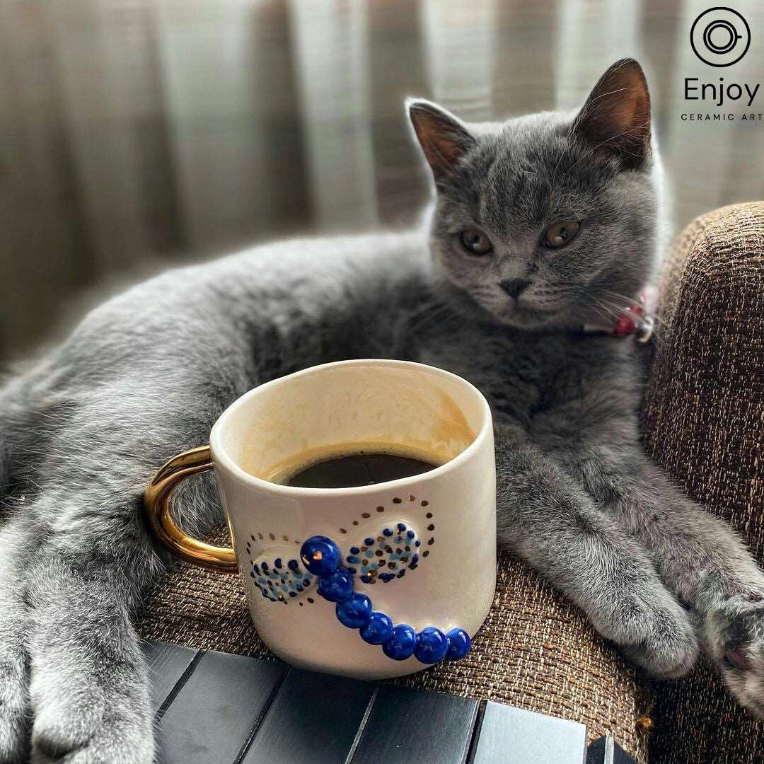 A serene grey kitten lounging next to a 'Blue Dragonfly Handmade Ceramic Coffee Mug' with gold handle, filled with coffee, symbolizing comfort and warmth in a home setting.