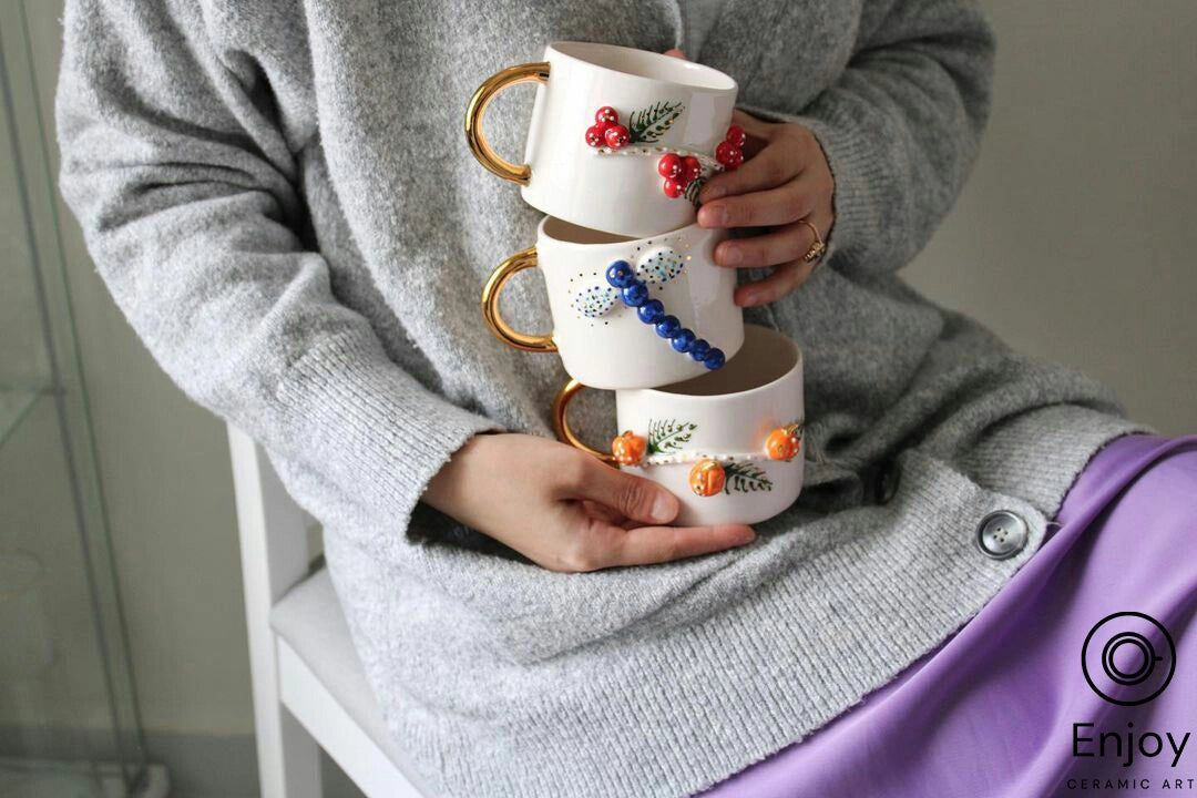 A person holding a stack of three handmade ceramic coffee mugs with gold handles, each adorned with a different colorful beadwork design including a blue dragonfly, red berries, and orange pumpkins, representing a collection of artisan-crafted mugs.