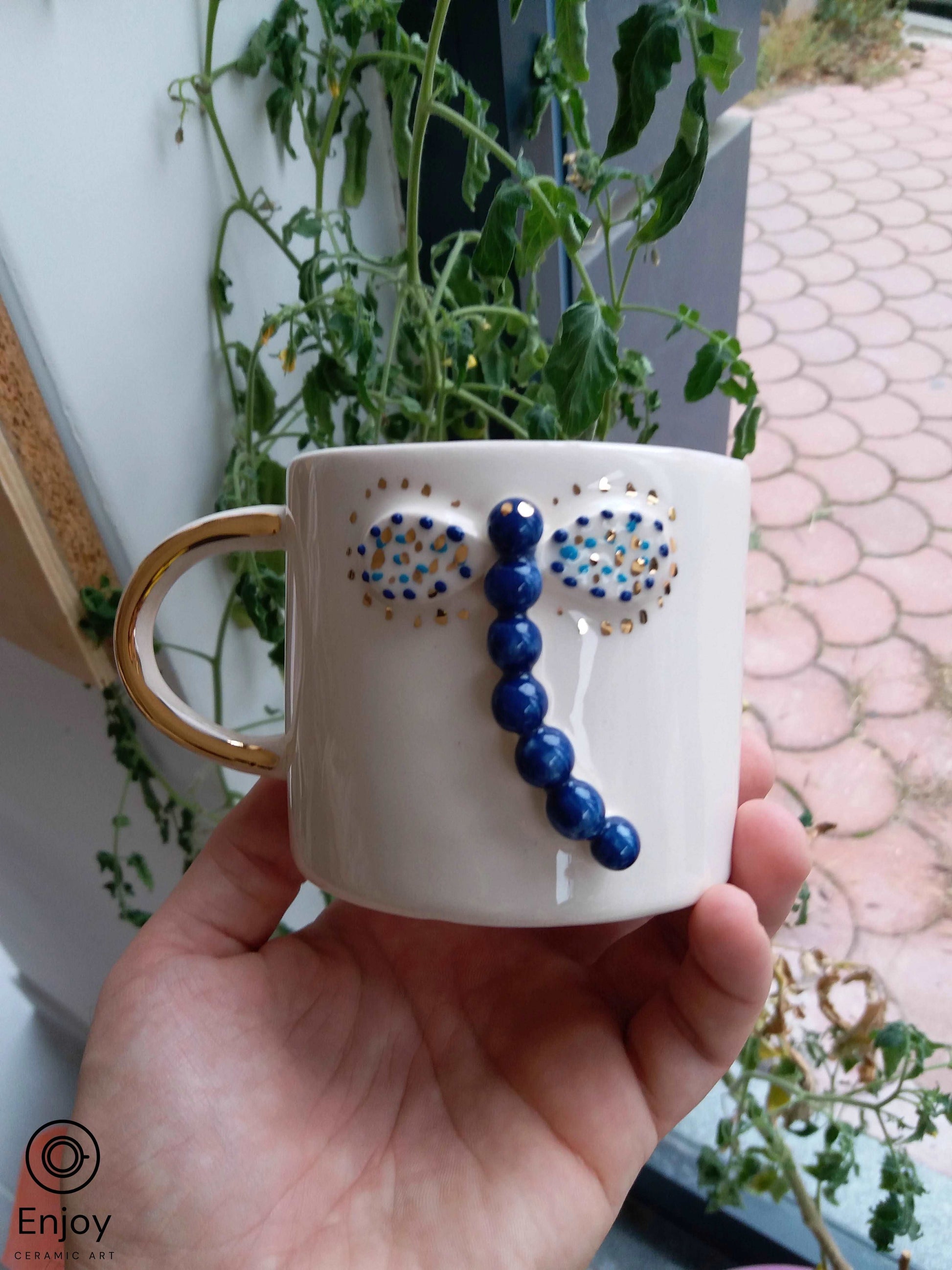 A hand presents a 'Blue Dragonfly Handmade Ceramic Coffee Mug' with a gold handle, set against a natural backdrop of green tomato plants and a tiled patio.