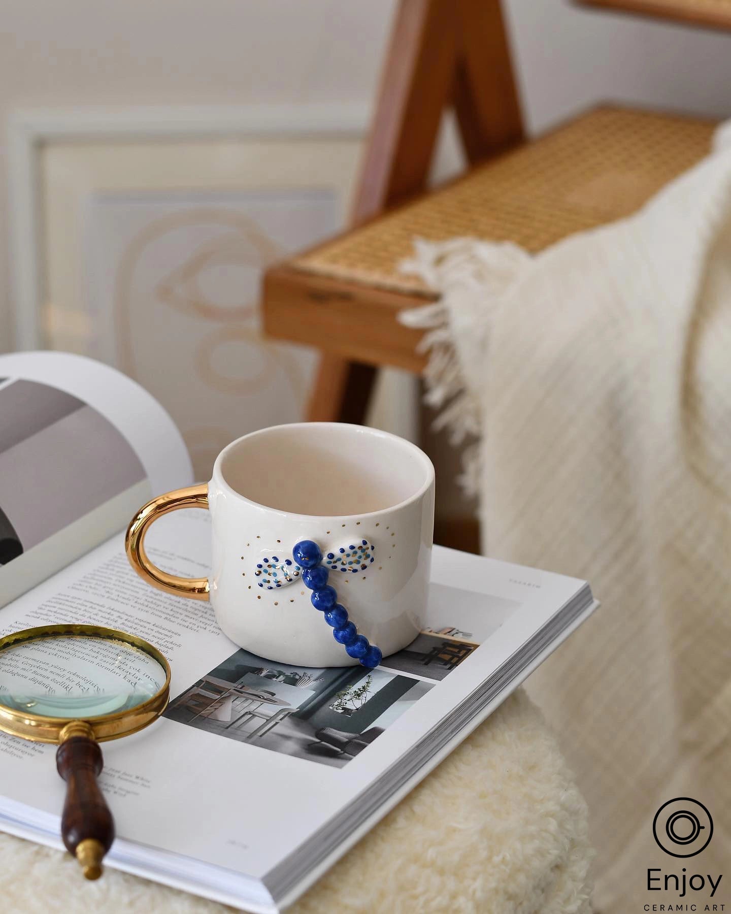 Cozy reading corner featuring the 'Blue Dragonfly Handmade Ceramic Coffee Mug, 10oz,' artisan-crafted with a unique blue beaded dragonfly design and a luxurious gold handle, resting on an open book beside a vintage magnifying glass.