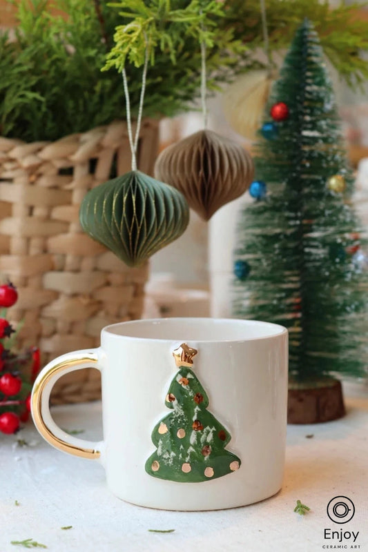 A festive white mug with a golden handle, adorned with a textured Christmas tree design in green, set against a backdrop of seasonal decorations.