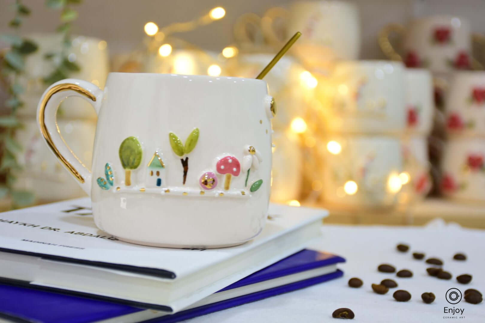 White ceramic mug with a whimsical garden design, featuring trees, a mushroom, and a small house. Set on stacked books with coffee beans scattered nearby.