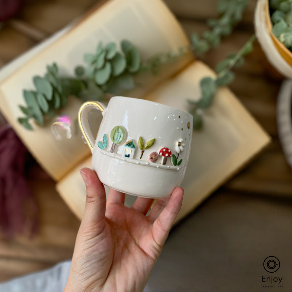 A hand holds a whimsical white mug featuring a 3D forest scene with trees, mushrooms, and a small house. Open books and greenery are in the background.