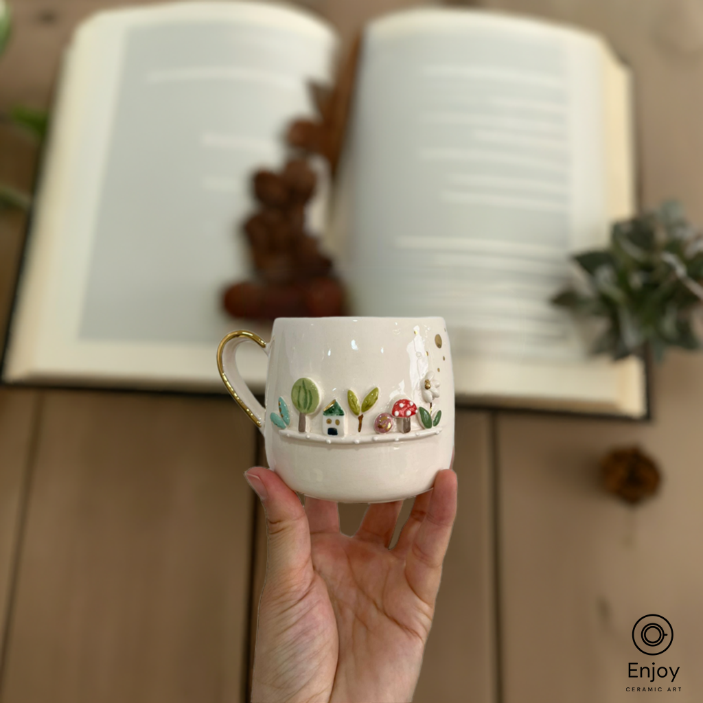 A white mug featuring a 3D garden scene with trees, a house, mushrooms, and a daisy, held in front of an open book.