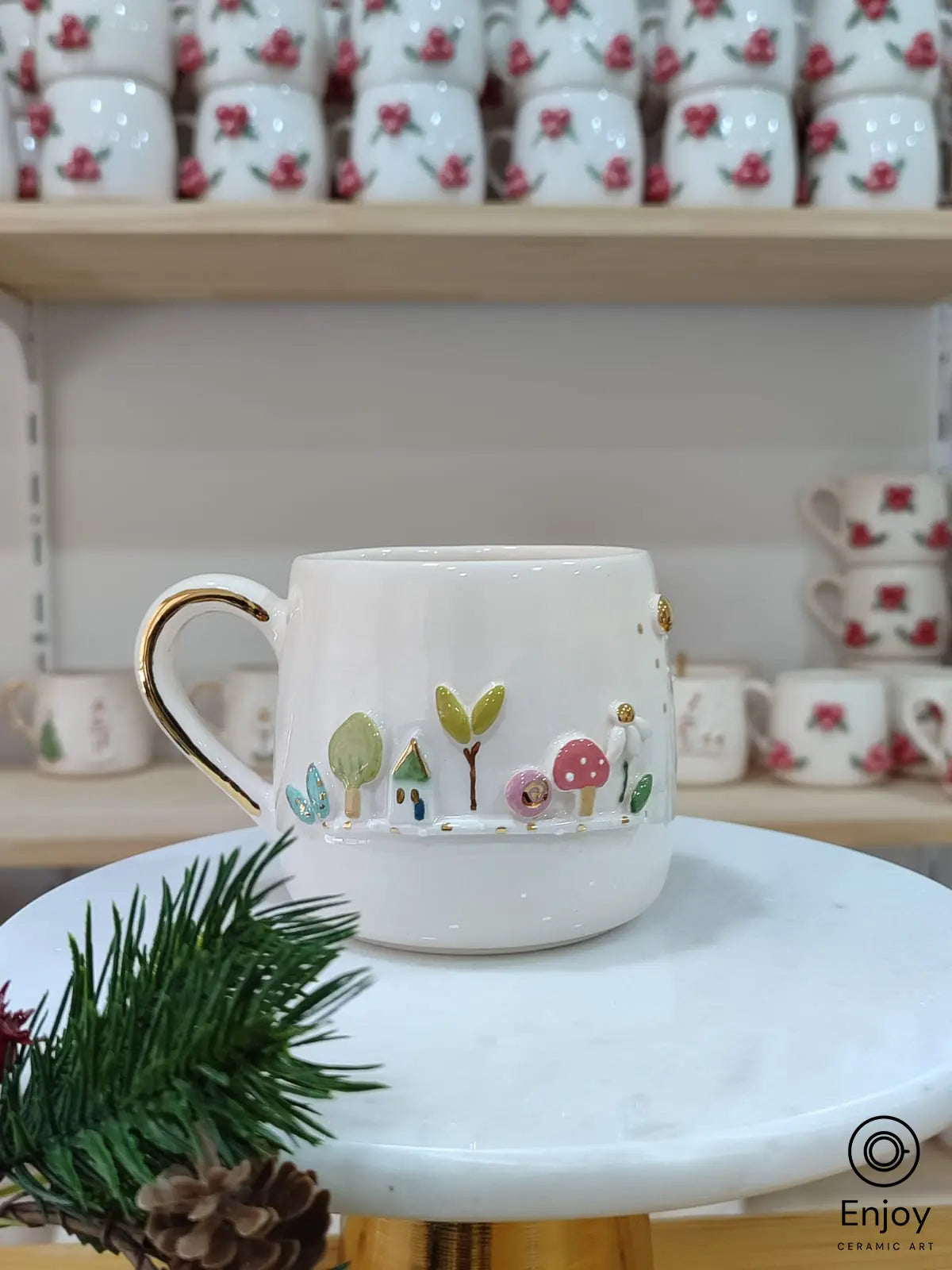 Handmade ceramic mug featuring a whimsical forest scene with trees, mushrooms, and a tiny house in vibrant colors, accented with a golden-rimmed handle. Displayed on a marble stand with festive touches, this mug brings a magical woodland charm to your everyday coffee or tea moments.