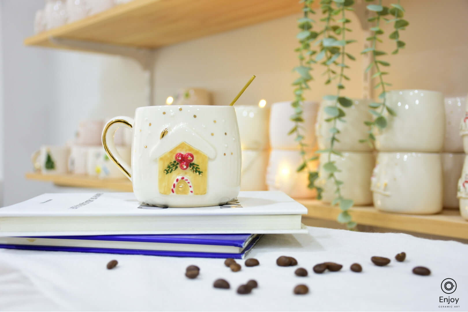 White ceramic mug featuring a festive gingerbread house design with red berries and green accents. Set on stacked books with coffee beans scattered nearby.