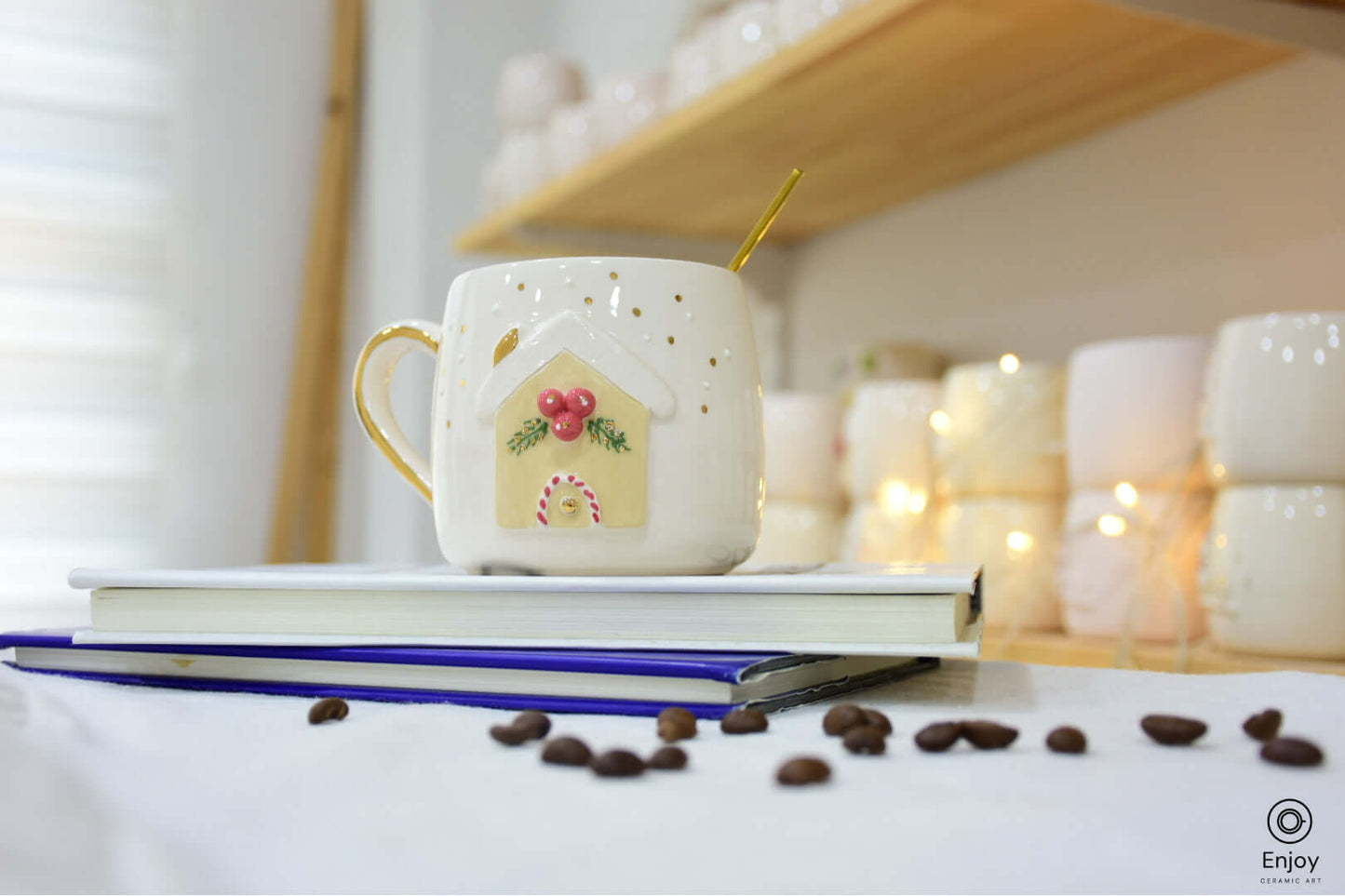 Festive white ceramic mug with a gingerbread house design, red berry accents, and a golden handle. Set on books with coffee beans scattered around.