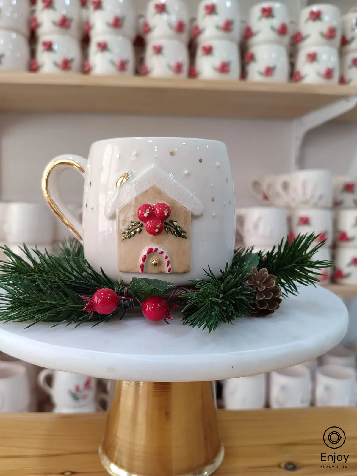 Handmade ceramic mug featuring a gingerbread house design with festive red berries, displayed on a marble stand with pine and pinecone accents, bringing a cozy holiday feel.