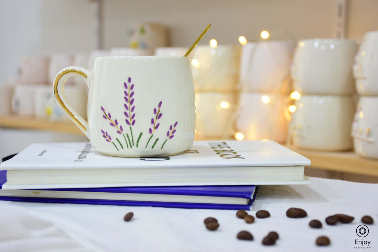 A lavender-themed ceramic mug with a golden handle sits on stacked books, surrounded by scattered coffee beans and soft fairy lights in the background.