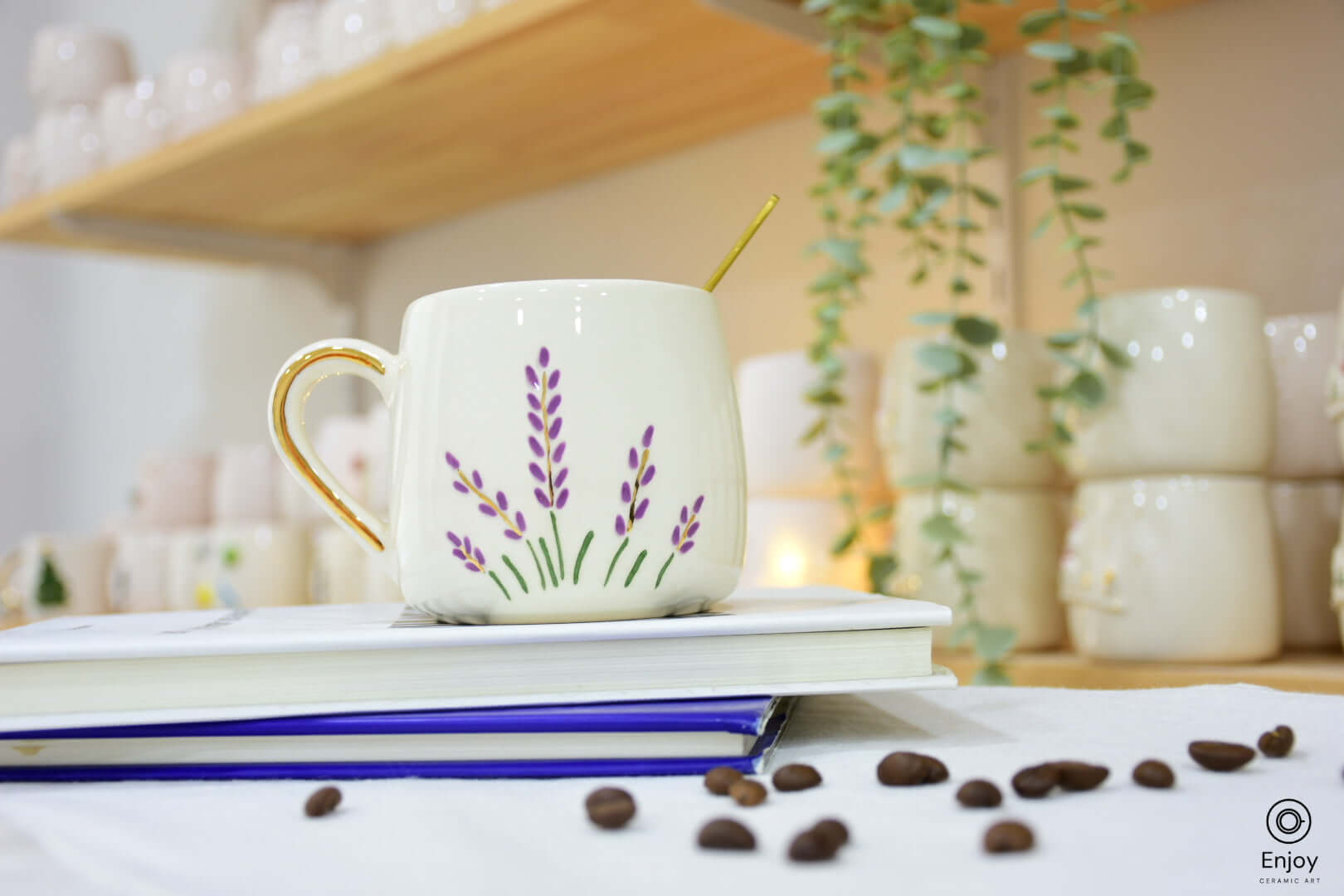 A ceramic mug with lavender designs and a gold handle sits on stacked books, surrounded by scattered coffee beans, with soft lighting and greenery in the background.