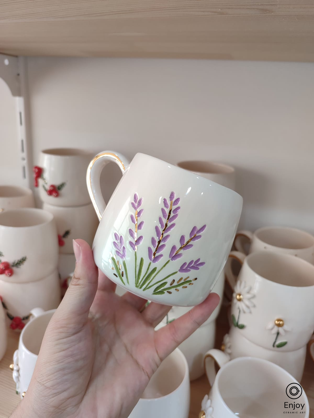 Handmade ceramic mug with lavender design and gold handle, held in front of a ceramic shelf.