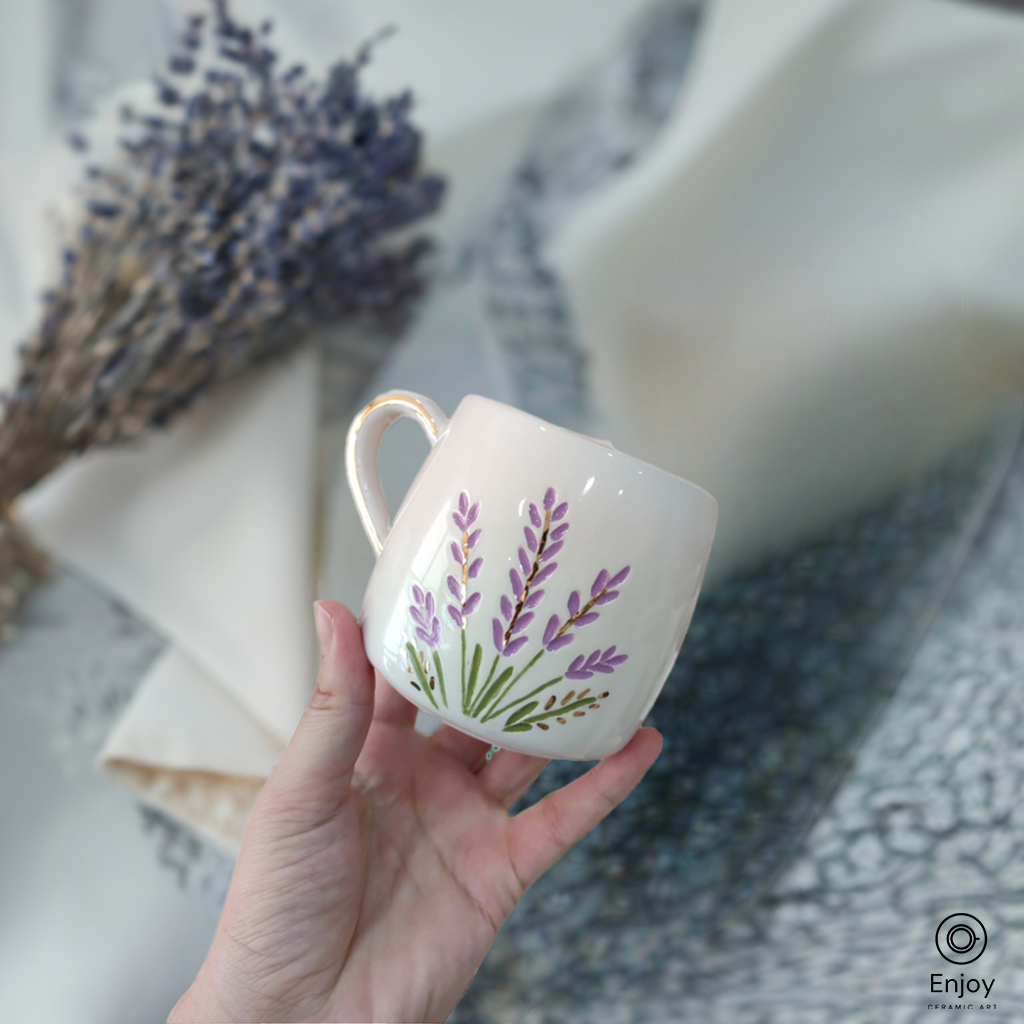 Handmade ceramic mug with lavender design and gold handle, held against a soft lavender backdrop.