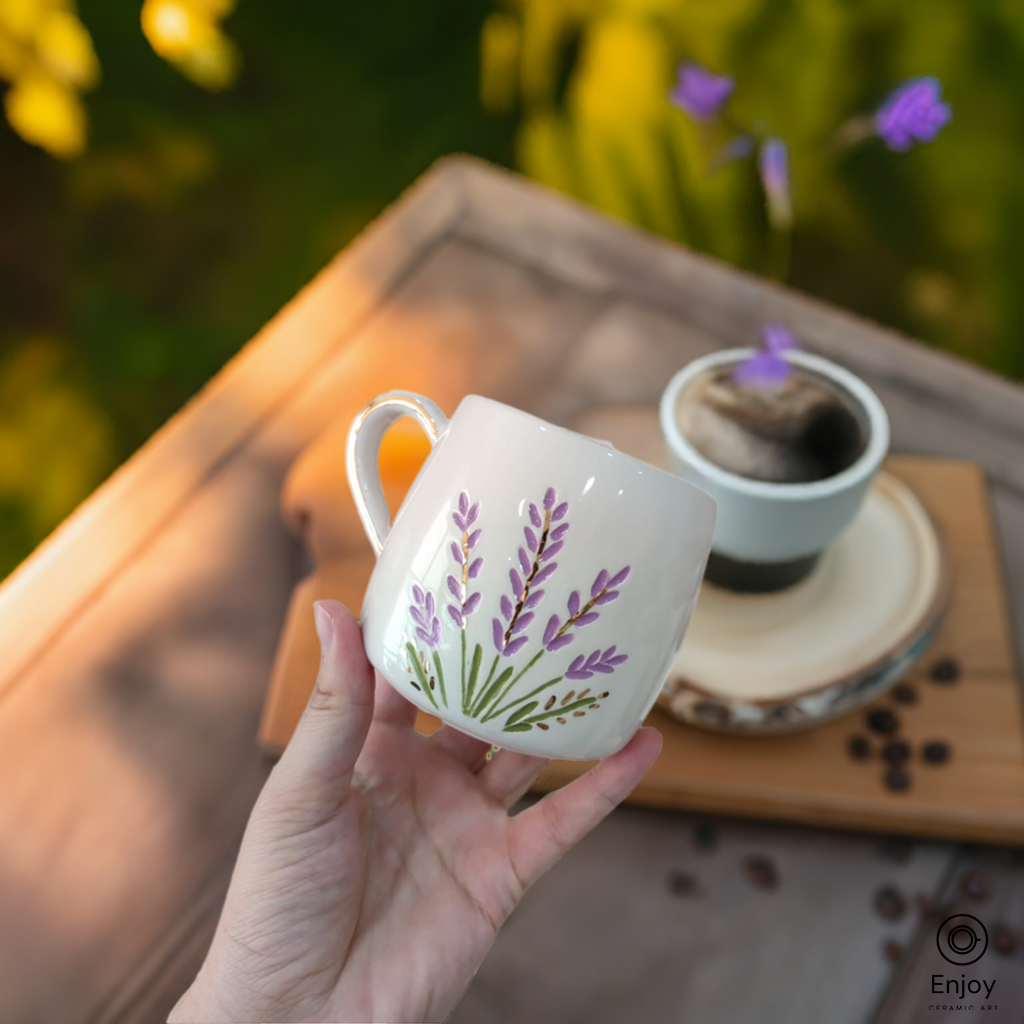 Handcrafted lavender-themed ceramic mug with a gold handle, held in a garden setting next to coffee.