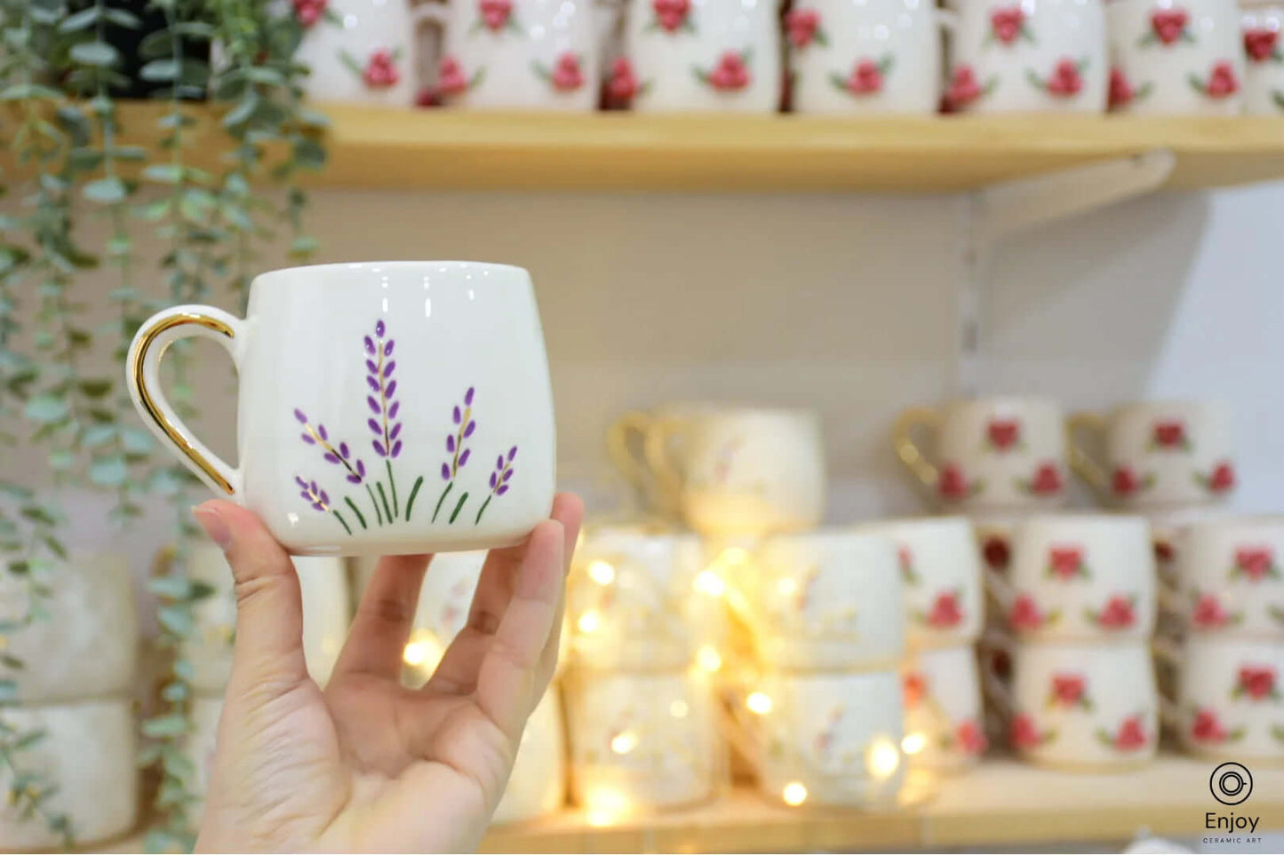 A hand holding a lavender-themed ceramic mug with a gold handle, against a backdrop of stacked floral mugs and soft, warm fairy lights.