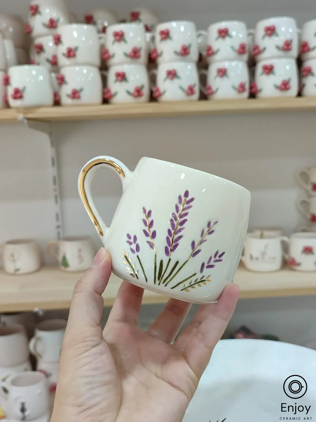 Handmade ceramic mug featuring delicate lavender stems painted in purple and gold accents, complemented by a gold-rimmed handle. Displayed against a backdrop of stacked floral mugs on wooden shelves.