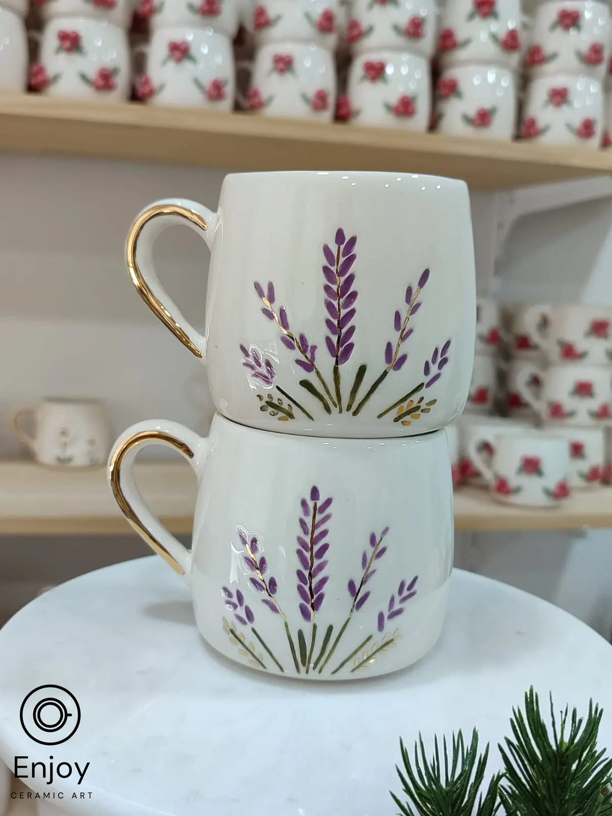 Stacked handmade ceramic mugs adorned with lavender stems in purple and gold accents, featuring gold-rimmed handles. Displayed elegantly in a cozy setting with floral-themed mugs on wooden shelves. Perfect for adding a touch of nature to your coffee moments.