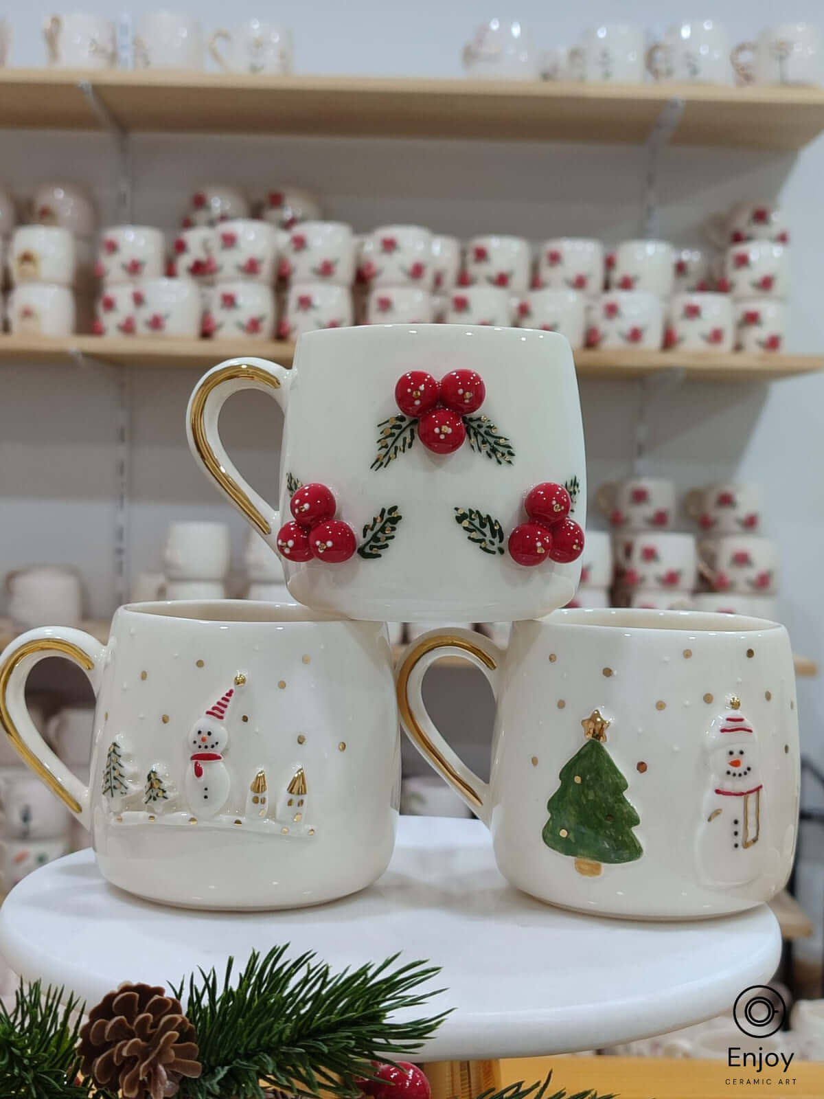 Set of three handmade Christmas-themed ceramic mugs featuring snowman, Christmas tree, and winter berries designs with gold handles.