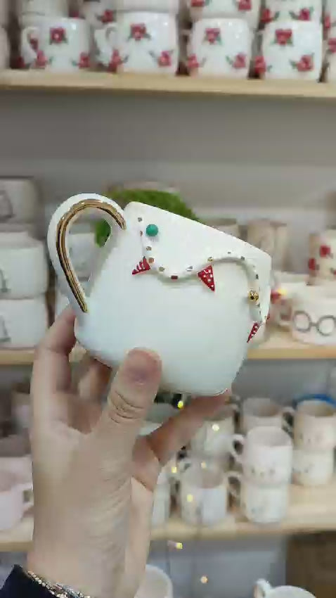 A close-up video showcasing a festive ceramic mug adorned with a 3D garland design, complete with tiny red and green bunting and gold accents. The mug has a shiny gold handle, sitting on a cozy holiday-themed background with fairy lights softly glowing in the distance, creating a warm and cheerful holiday vibe.