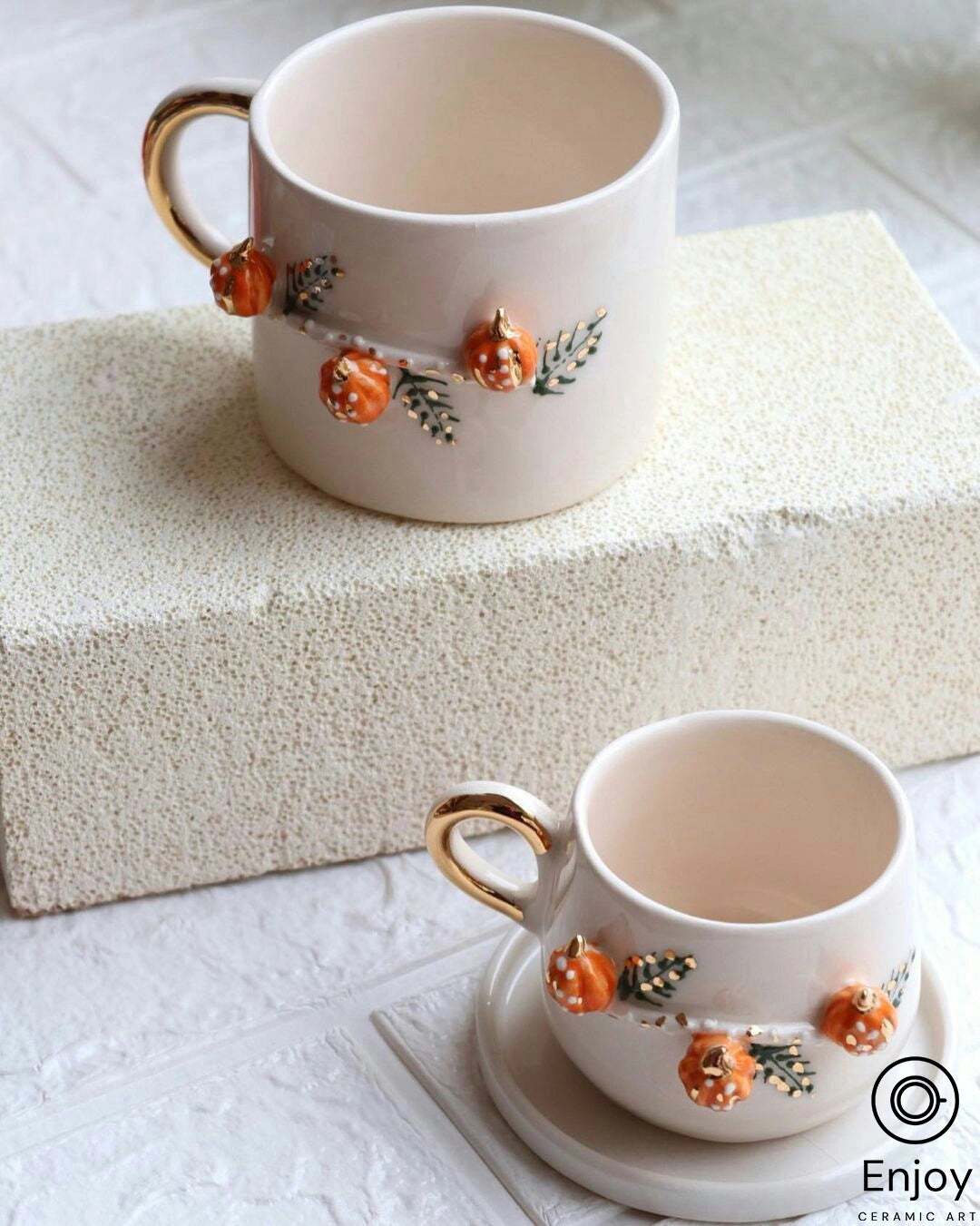 Two handcrafted ceramic mugs with golden handles and autumn-themed pumpkin designs, set on a creamy textured surface for an elegant, seasonal display.