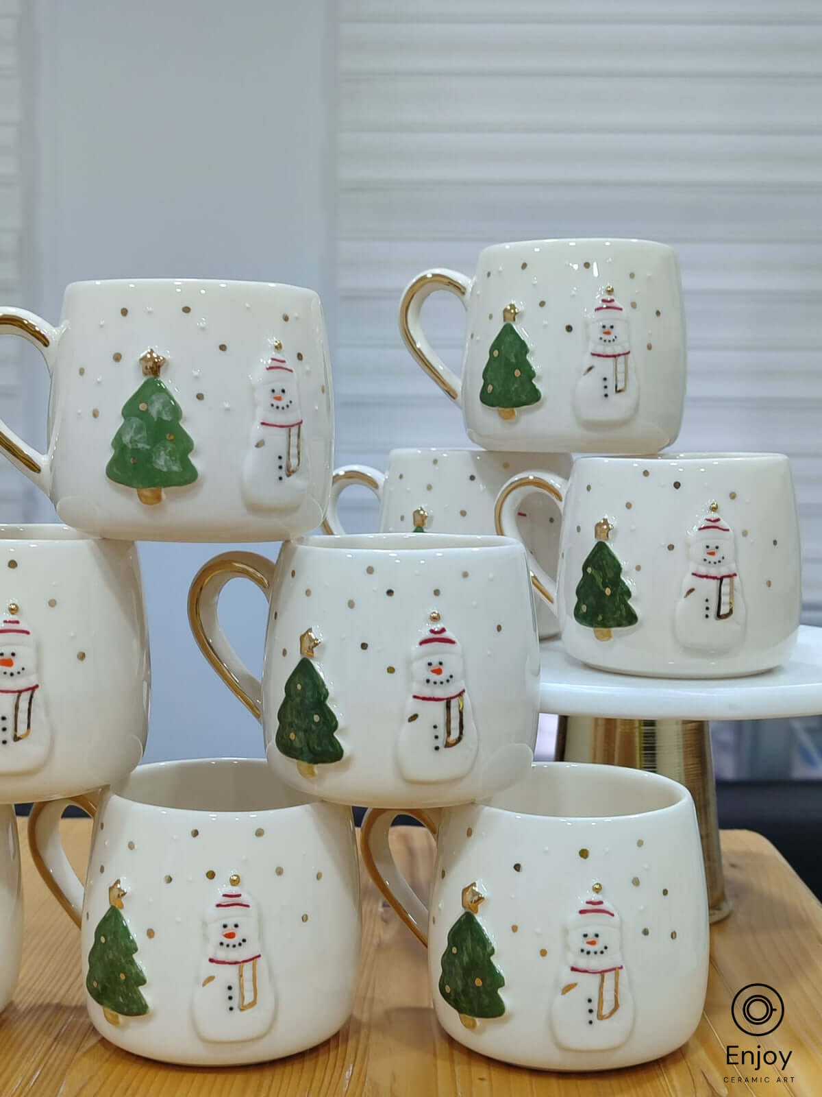 A charming collection of handmade ceramic mugs adorned with festive snowman and Christmas tree motifs, each delicately highlighted with golden accents. These mugs bring warmth and joy to any holiday gathering, perfect for enjoying a cozy cup of cocoa or gifting to loved ones during the season.