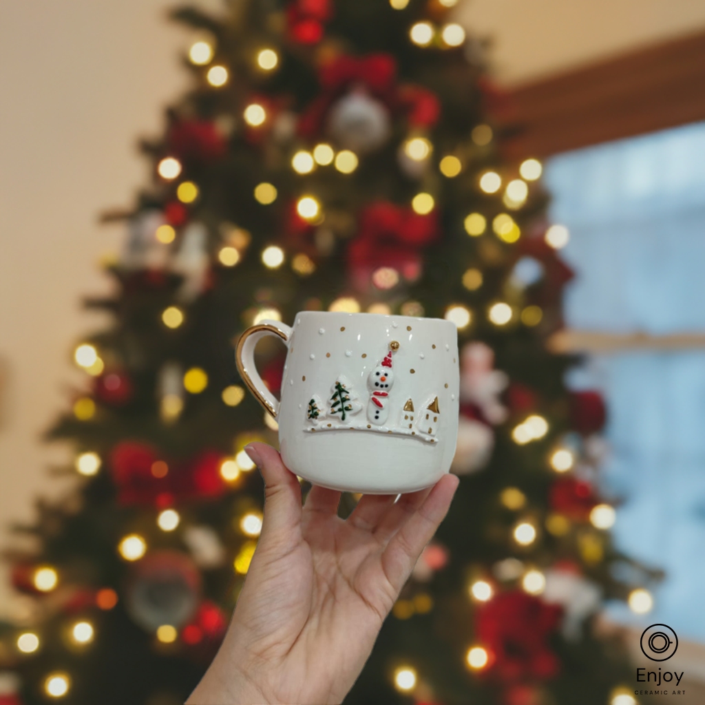 Hand holding a snowman-themed ceramic mug with a golden handle, in front of a glowing Christmas tree.