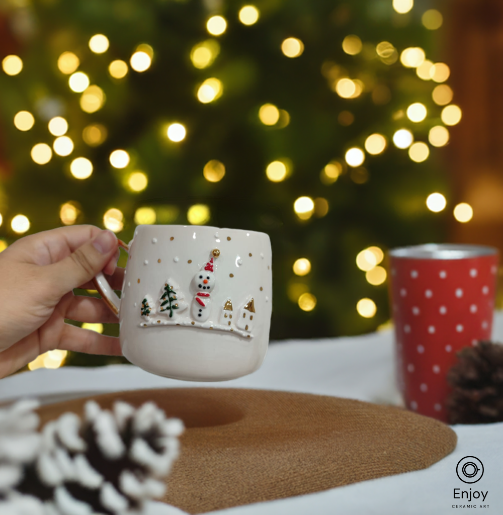 Hand holding a festive ceramic mug with a snowman and Christmas tree design, in front of twinkling holiday lights.