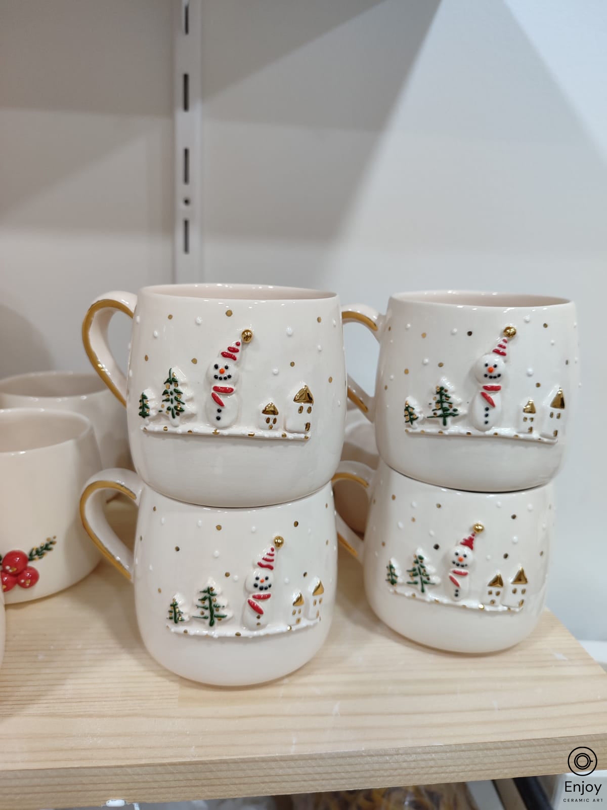 A set of four handmade ceramic snowman mugs with festive gold details, displayed on a wooden shelf.