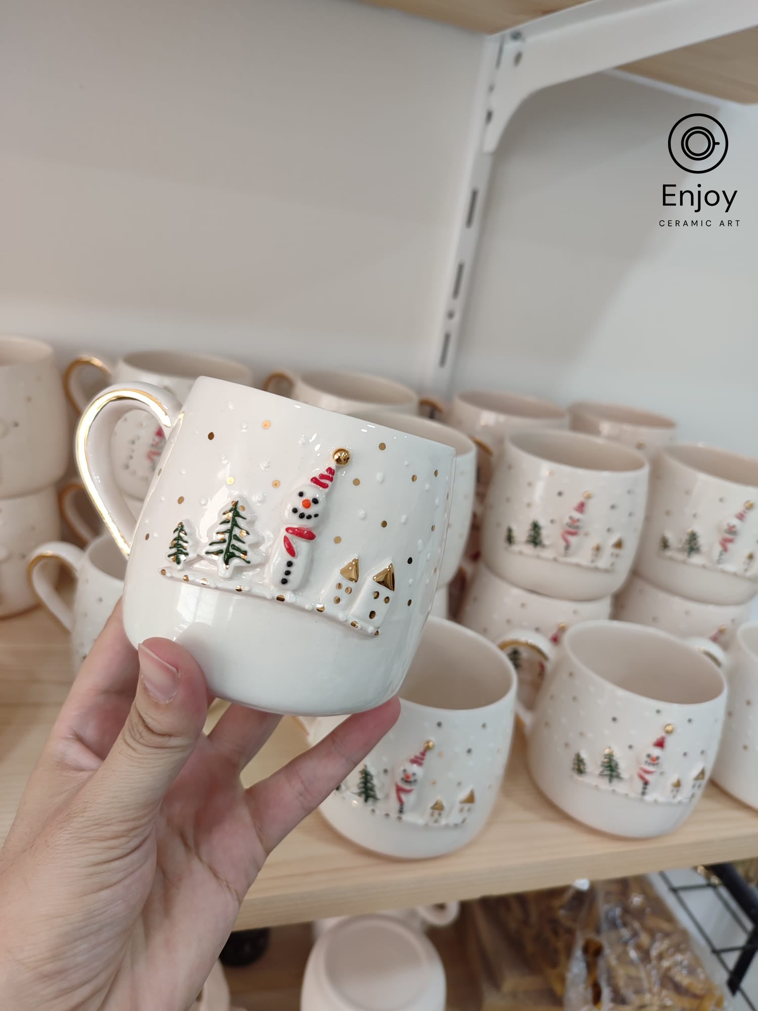 A hand holding a handmade ceramic mug featuring a snowman and festive village scene, with gold accents, against a shelf filled with similar Christmas-themed mugs.