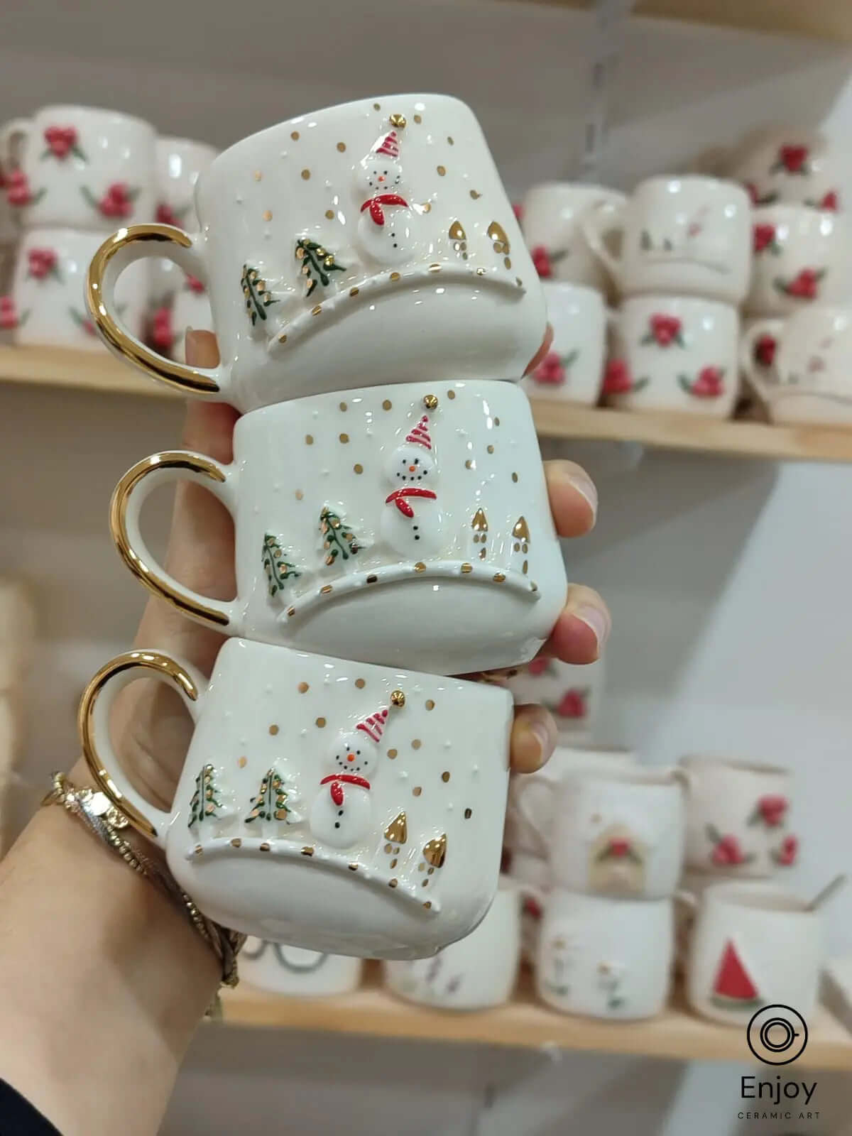 A trio of festive snowman espresso cups with gold handles, showcasing intricate holiday designs, held in front of a mug display shelf.