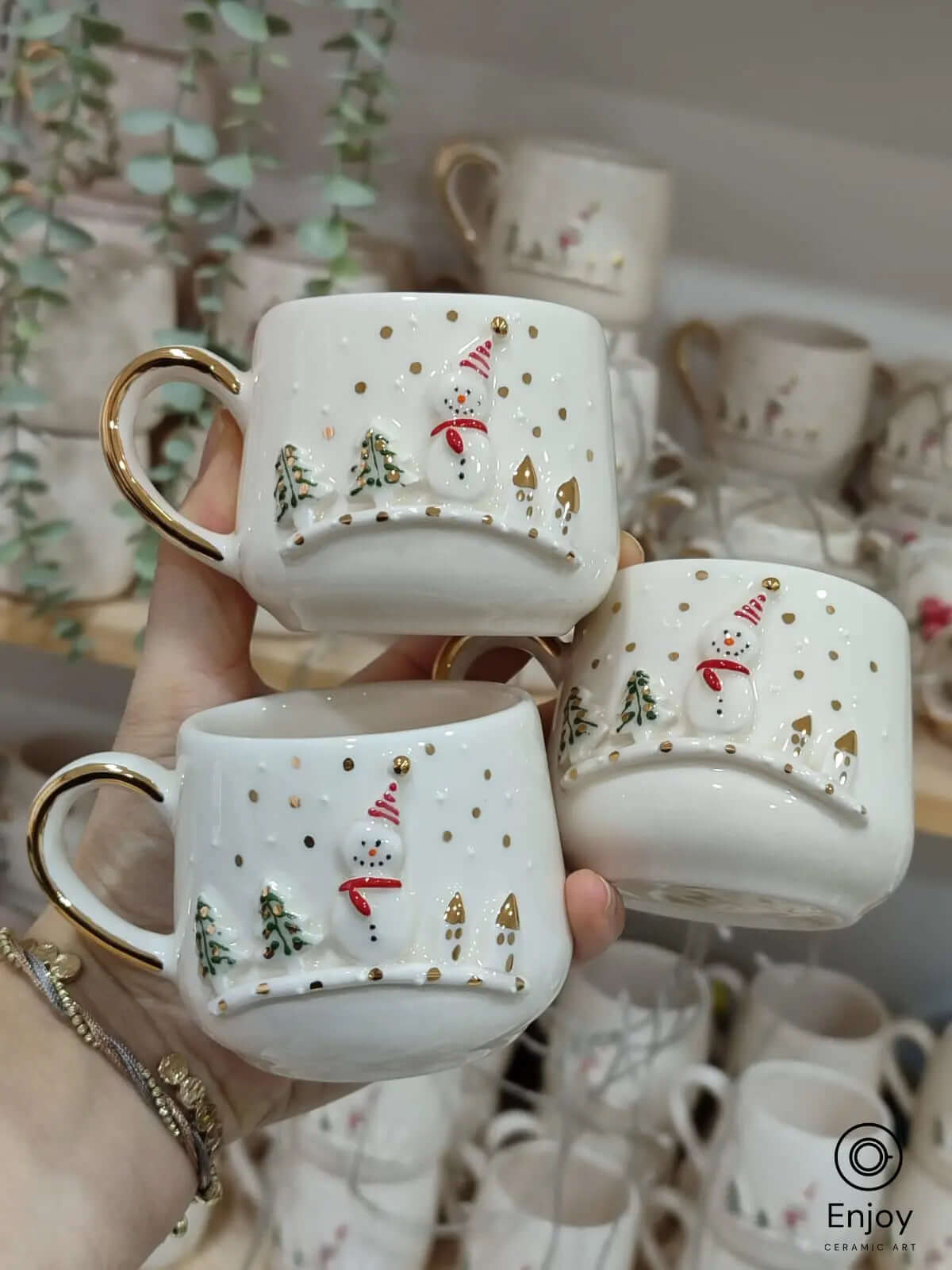 Three handcrafted snowman espresso cups with gold accents, featuring festive designs of snowmen, trees, and gold dots, held against a mug display.