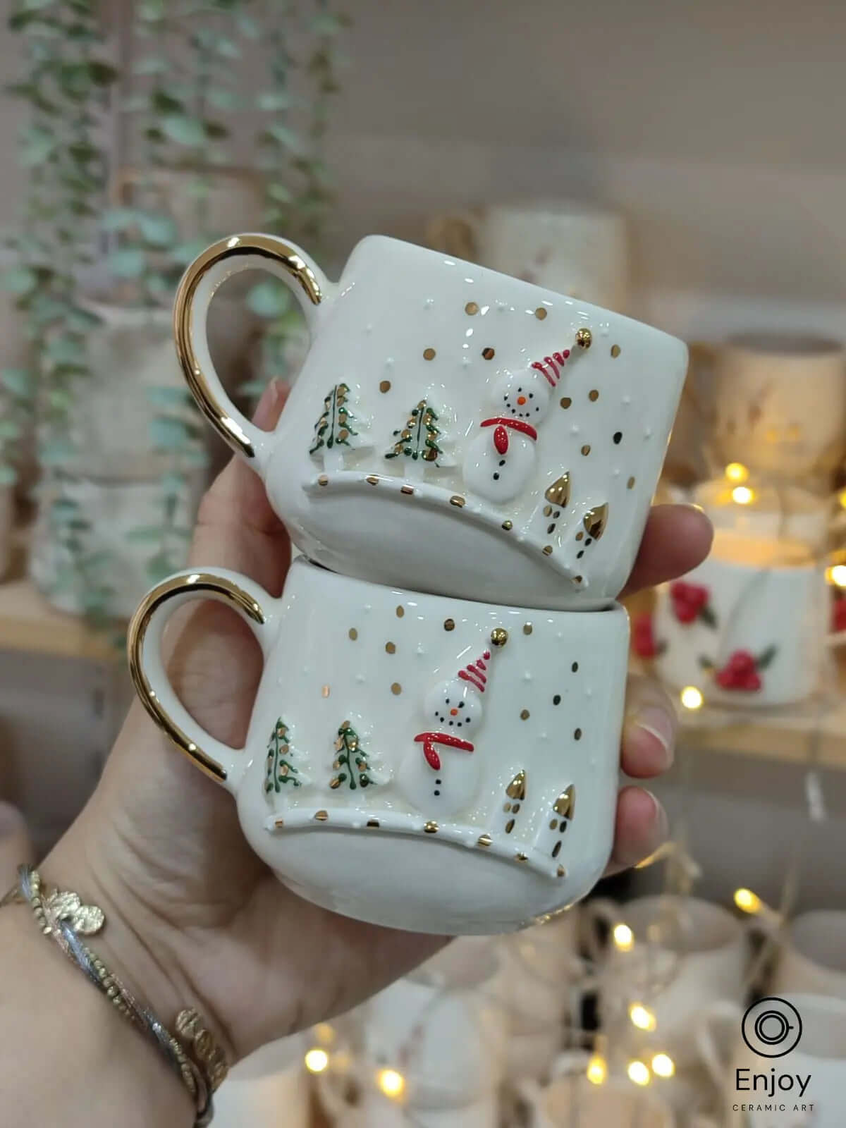 Pair of snowman-themed espresso cups with gold handles, held against a festive backdrop of fairy lights and ceramic mug shelves.