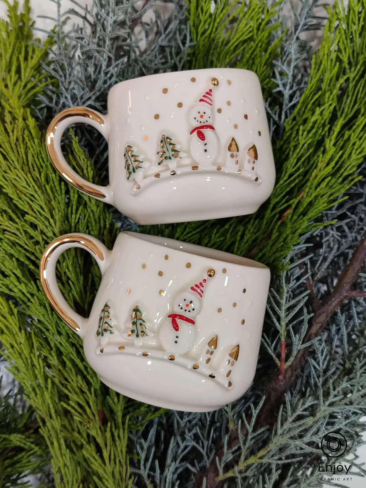 Set of two snowman-themed espresso cups with gold accents, placed on lush greenery for a festive and elegant holiday presentation.