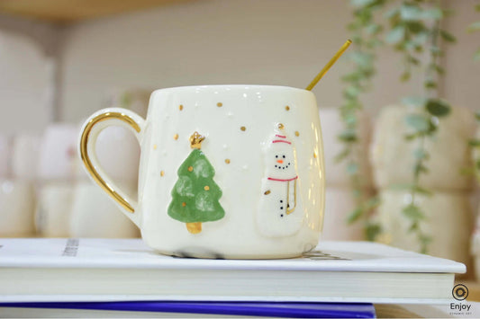 A festive ceramic mug featuring a snowman and Christmas tree design, accented with gold dots and a golden handle, perfect for holiday cheer.