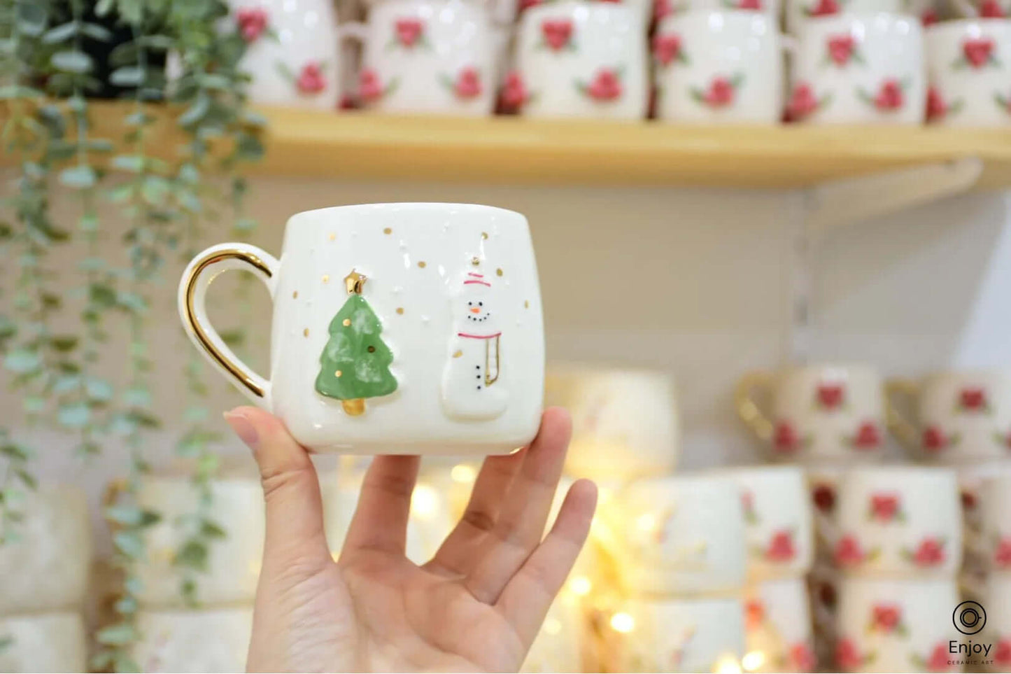 Hand holding a festive ceramic mug decorated with a green Christmas tree, a snowman, and gold accents, perfect for holiday cheer.