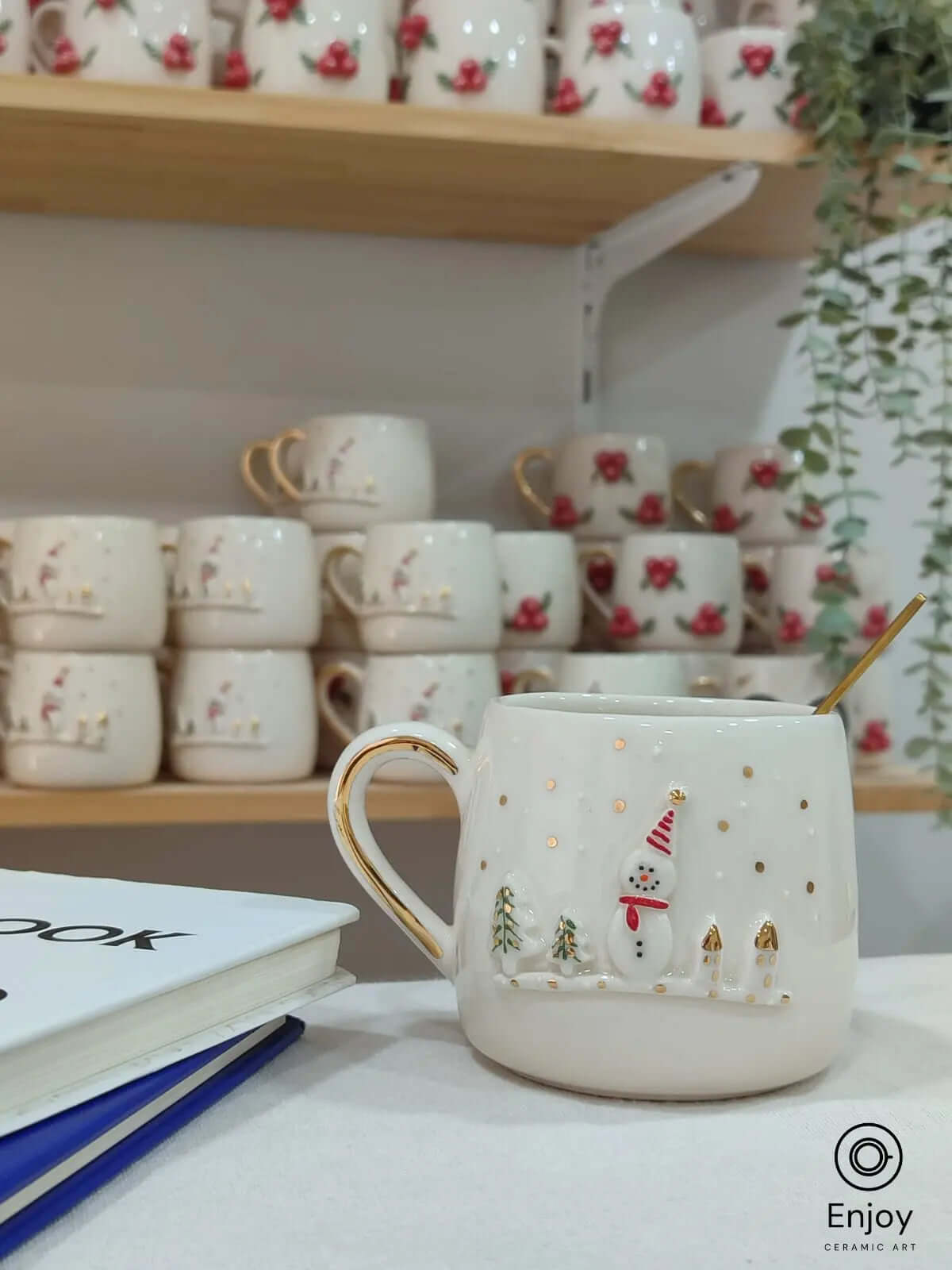 A charming handmade ceramic mug adorned with a joyful snowman, golden accents, and festive trees. This cozy piece sits perfectly beside a notebook, creating the ideal winter setup. Whether you're journaling or planning your holiday season, this delightful mug brings warmth and seasonal spirit to your moments.