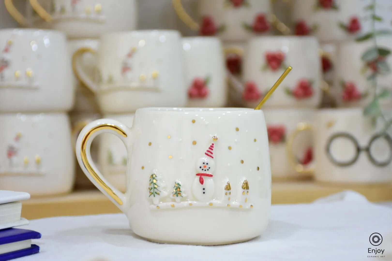 A festive ceramic mug featuring a snowman and gold-accented trees, with a golden handle, surrounded by other holiday-themed mugs in the background.