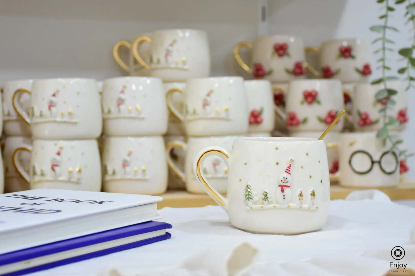 A ceramic mug featuring a festive snowman and golden trees, with a gold-accented handle, placed in front of other holiday-themed mugs on a shelf.